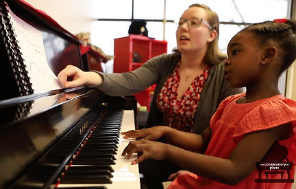 A young girl playing piano while seated next to her teacher