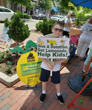 A piano lesson student holding a sign that reads "give a donation. get a lemonade. help kids"
