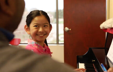 A young girl smiling up at her piano instructor while seated in front of a piano